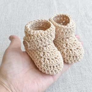 Pode incluir: Um par de sapatinhos de beb&eacute; em croch&ecirc; bege com um bot&atilde;o em um sapatinho.