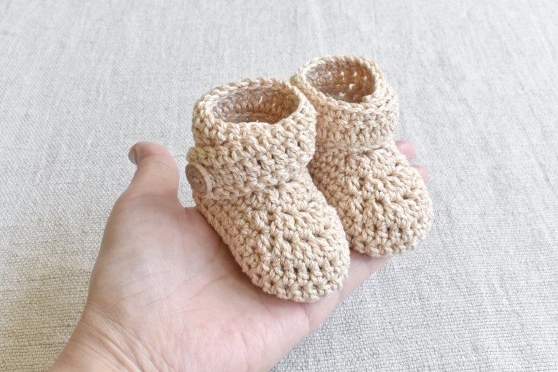 Pode incluir: Um par de sapatinhos de beb&eacute; em croch&ecirc; bege com um bot&atilde;o em um sapatinho.
