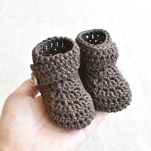 Pode incluir: Um par de botinhas de beb&eacute; em croch&ecirc; castanho com um bot&atilde;o em uma botinha.
