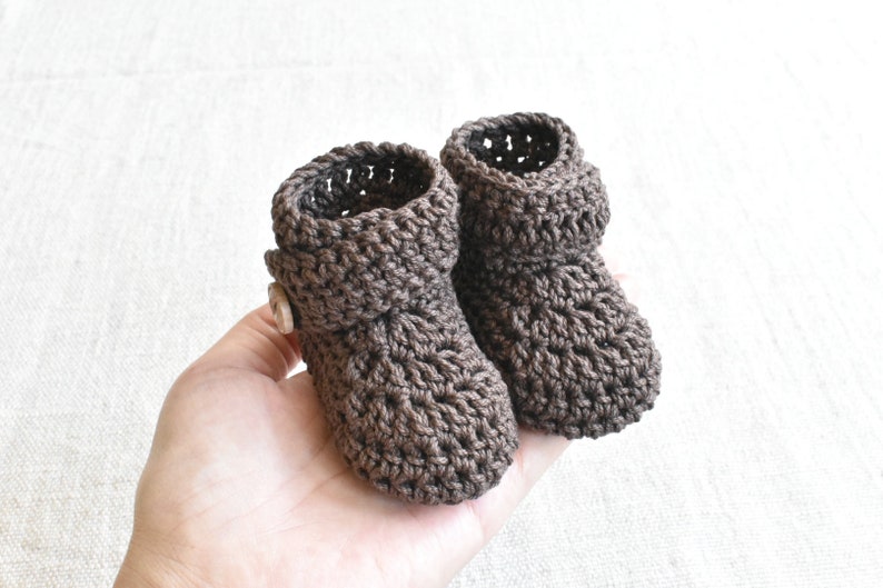 Pode incluir: Um par de botinhas de beb&eacute; em croch&ecirc; castanho com um bot&atilde;o em uma botinha.
