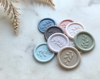 May include: A collection of round wax seals in various pastel colors, including blue, pink, and green. Each seal features a raised botanical design of a leafy branch. The seals are arranged on a white marble surface, with a sprig of dried flowers in the background.