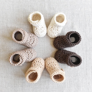 Pode incluir: Seis pares de sapatinhos de beb&eacute; em croch&ecirc; em v&aacute;rios tons de castanho e branco. Os sapatinhos est&atilde;o dispostos em c&iacute;rculo sobre uma superf&iacute;cie texturizada cinzenta clara.