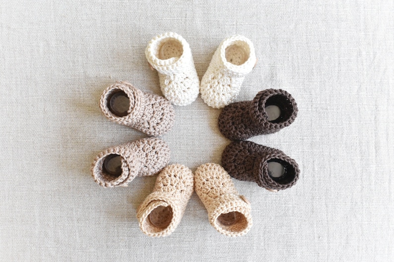 Pode incluir: Seis pares de sapatinhos de beb&eacute; em croch&ecirc; em v&aacute;rios tons de castanho e branco. Os sapatinhos est&atilde;o dispostos em c&iacute;rculo sobre uma superf&iacute;cie texturizada cinzenta clara.