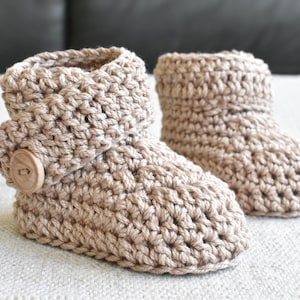 Pode incluir: Um par de botinhas de beb&eacute; em croch&ecirc; bege com um bot&atilde;o de madeira em uma botinha.