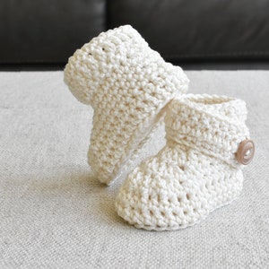 Pode incluir: Um par de sapatinhos de beb&eacute; de crochet branco com um bot&atilde;o castanho em um sapatinho.
