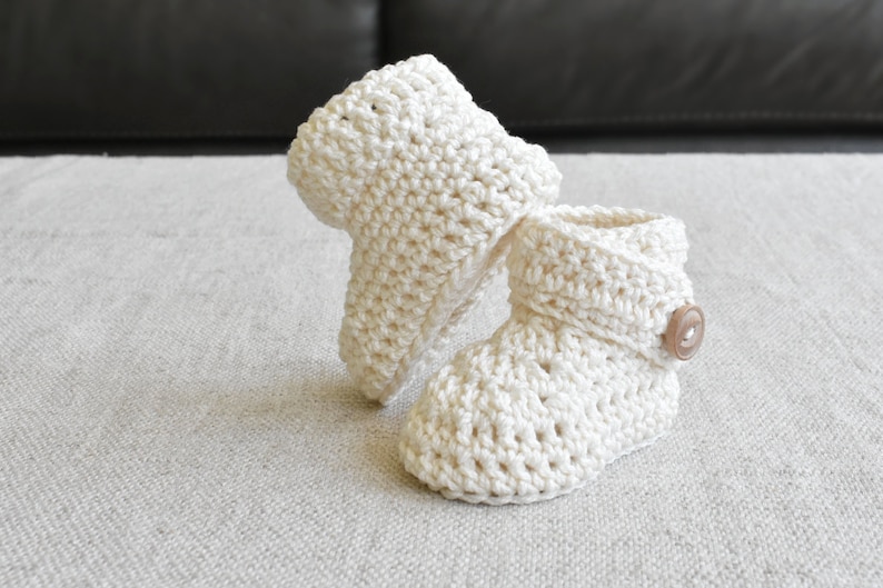 Pode incluir: Um par de sapatinhos de beb&eacute; de crochet branco com um bot&atilde;o castanho em um sapatinho.