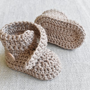 Pode incluir: Um par de sapatinhos de beb&eacute; de croch&ecirc; em cor castanho claro. Os sapatinhos t&ecirc;m um fecho de bot&atilde;o e s&atilde;o feitos com um fio macio e texturizado.