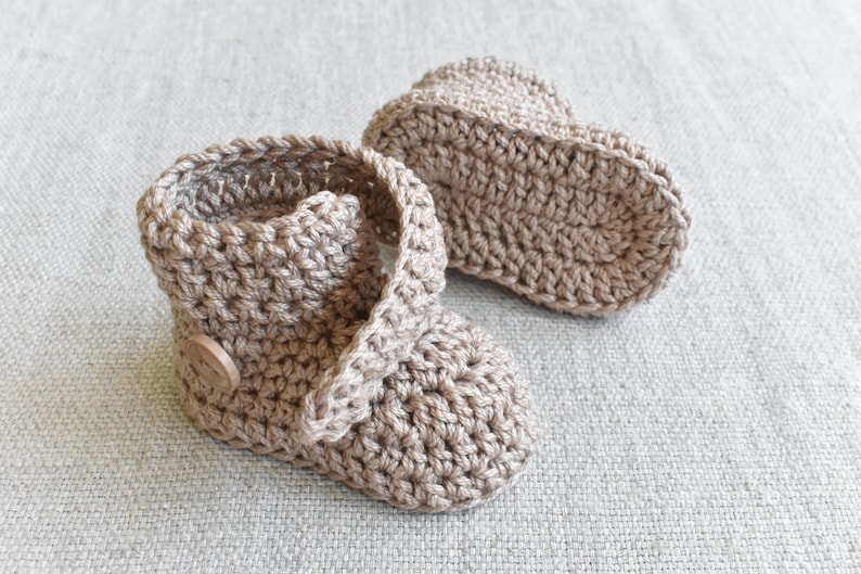 Pode incluir: Um par de sapatinhos de beb&eacute; de croch&ecirc; em cor castanho claro. Os sapatinhos t&ecirc;m um fecho de bot&atilde;o e s&atilde;o feitos com um fio macio e texturizado.