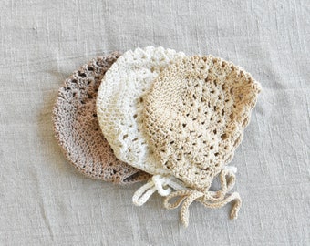 May include: Three crocheted baby bonnets in beige, white, and brown. The bonnets have a delicate lace pattern and are tied with matching ribbons.