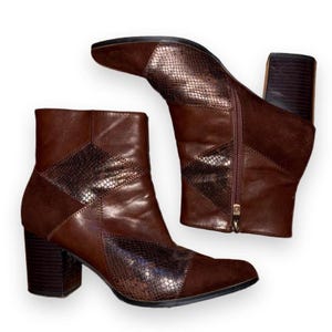 May include: A pair of brown ankle boots featuring a block heel, a side zipper closure, and a combination of smooth leather, suede, and metallic snakeskin-textured panels. The boots have a pointed toe and a dark brown stacked heel design.