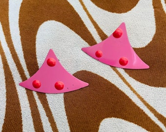 May include: Two pink, triangular earrings with three red, circular accents each. The earrings are displayed on a brown and white patterned fabric, creating a retro aesthetic. These earrings are a fun, colorful accessory.