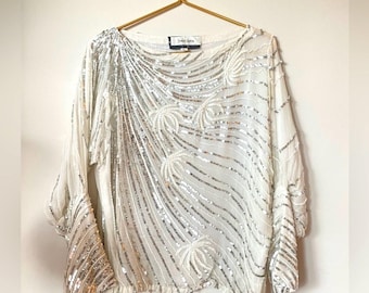 May include: A white, long-sleeved blouse with a scoop neck and silver sequins in a wavy pattern. The blouse has a relaxed fit and is made of a lightweight fabric.