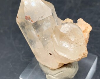 Puede incluir: Un grupo de cristales de cuarzo claros y translúcidos con una terminación puntiaguda y un cristal secundario más pequeño unido al lado. La base del espécimen mineral está anclada en un soporte de masilla gris texturizada sobre una base cuadrada de acrílico transparente.