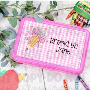 May include: A pink lunchbox with a gingham pattern and the name "Brooklyn Jane" printed on the lid. The lid features a pencil graphic with a pink bow. Crayons are visible in the background.