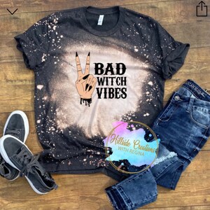 Puede incluir: Una camiseta negra con un diseño decolorado y las palabras "BAD WITCH VIBES" en negro. La camiseta presenta una mano haciendo el signo de la paz con uñas largas y negras. Vaqueros oscuros y zapatillas grises completan el conjunto.