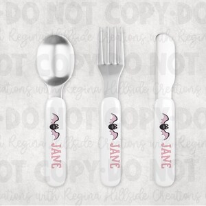 May include: A three-piece children's flatware set featuring a spoon, fork, and butter knife. Each piece has a white handle with a pink bat design and the name "JANE" printed in pink. The utensils are made of stainless steel.