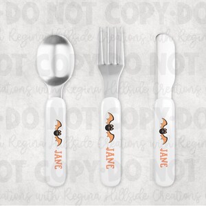 May include: A three-piece Halloween-themed flatware set. The set includes a spoon, fork, and knife. Each piece has a white handle with an orange bat design and the name "JANE" printed in orange. The utensils are made of shiny silver metal.