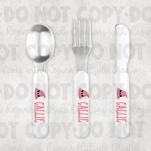 May include: A set of three personalized children's utensils: a spoon, fork, and knife. Each utensil has a white handle with a pink Santa hat design and the name "CALLIE" in pink. The spoon, fork, and knife are made of stainless steel.