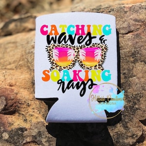 May include: White can cooler with the text "Catching waves & soaking rays" in colorful, retro-style lettering. The design includes sunglasses with a sunset scene and a leopard print frame. A logo is visible at the bottom.