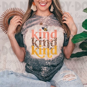 May include: A woman wearing a gray t-shirt with a bleached effect and a graphic print of a bee and the words "kind kind kind". The shirt has a pink, leopard print, and yellow design.