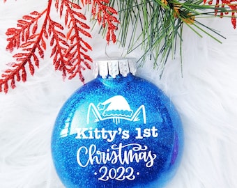 Puede incluir: Adorno navideño azul brillante con el texto "Kitty's 1st Christmas 2022". El adorno presenta un gato blanco con un gorro de Papá Noel. El follaje festivo rojo y verde está en el fondo.