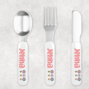 May include: A set of three personalized children's cutlery with white handles and pink floral designs. The spoon, fork, and knife are all labeled with the name "Jenna".