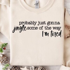 May include: Cream-colored sweatshirt with the text "probably just gonna jingle some of the way I'm tired" in black script. The sweatshirt is decorated with pinecones, greenery, and a wooden star. A cozy, casual top for the holiday season.
