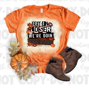 May include: Orange t-shirt with the text "GET IN LOSER WE'RE DOING FALL STUFF". The shirt is paired with brown ankle boots and fall-themed decorations, including pumpkins and leaves. A seasonal autumn design.