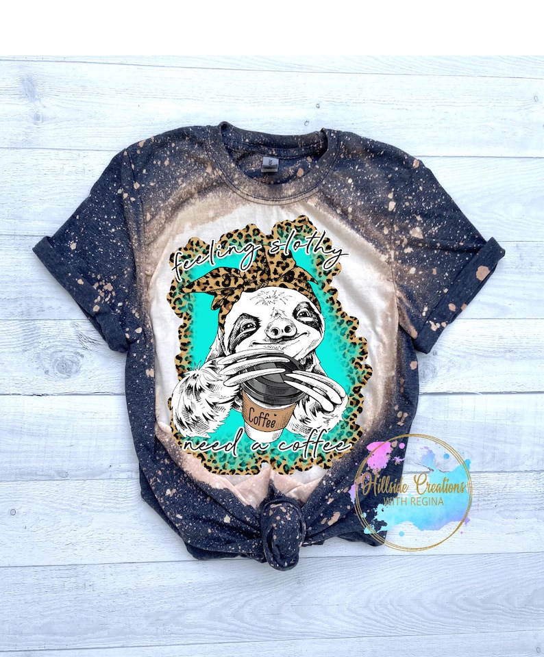 May include: A black and white graphic of a sloth wearing a leopard print headband and holding a coffee cup. The text "feeling sloth-y need a coffee" is printed on the shirt. The shirt is a black and white tie-dye.