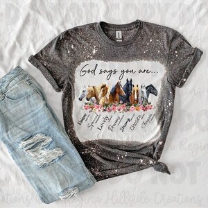May include: A gray t-shirt with a bleach-splattered design featuring a group of horses and the text "God says you are..." with words like "unique", "special", "lovely", "precious", "strong", "chosen", and "fearless" surrounding the horses.