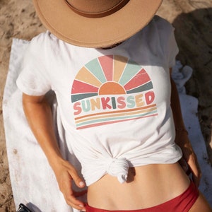 以下が含まれることがあります： レトロな太陽のグラフィックとカラフルな文字で「Sunkissed」という言葉が入った白いTシャツ。茶色の帽子と赤いビキニボトムを着用。黒いサングラスが白いタオルに置かれています。