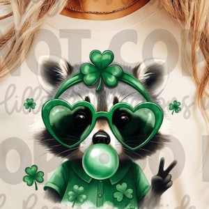 May include: A cream-colored t-shirt featuring a raccoon wearing heart-shaped green sunglasses and a headband with a shamrock. The raccoon is blowing a green bubble and giving a peace sign. Shamrock accents are scattered around the design.