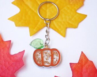 May include: A pumpkin-shaped keychain with an orange outline and gold flakes inside. It has a silver chain and ring, with a green leaf charm. The keychain is set against a white background with orange and red fall leaves.
