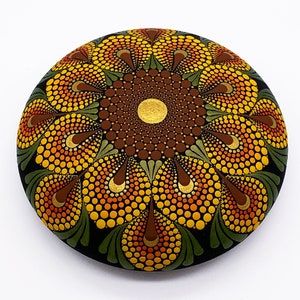 Könnte beinhalten: Ein runder Stein, der mit einem Mandala-Muster in Braun-, Orange-, Gelb- und Grüntönen bemalt ist. Das Mandala hat einen zentralen Kreis mit einem geometrischen Muster, umgeben von einer Reihe konzentrischer Kreise und Blütenblättern.