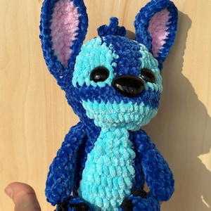 May include: A handmade plush toy in the shape of a blue alien character with large ears and black eyes. The toy is made of a soft, textured material with light blue and dark blue sections. The inside of the ears is pink.