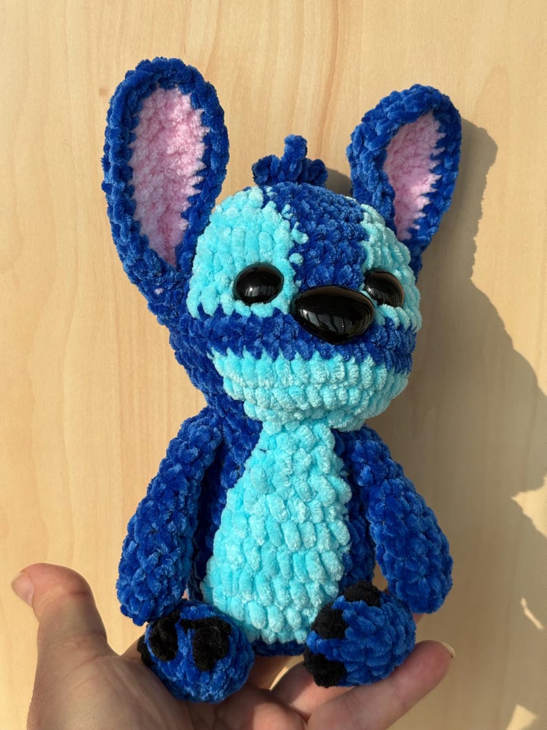 May include: A handmade plush toy in the shape of a blue alien character with large ears and black eyes. The toy is made of a soft, textured material with light blue and dark blue sections. The inside of the ears is pink.