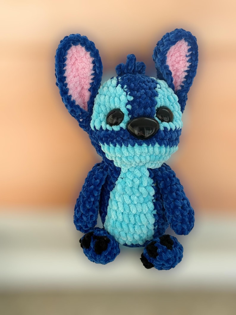 May include: A plush blue and light blue stuffed animal with large ears and black eyes. The stuffed animal has a textured, fuzzy appearance and is likely handmade. The ears have a pink interior. The toy is a character from a popular animated film.