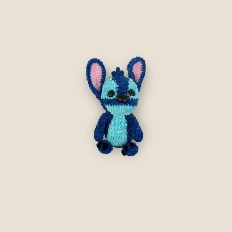 May include: A handmade crochet Stitch doll in shades of blue, with pink inner ears and black eyes. The plush toy features a detailed design, capturing the character's distinctive features. The doll is a charming collectible item.