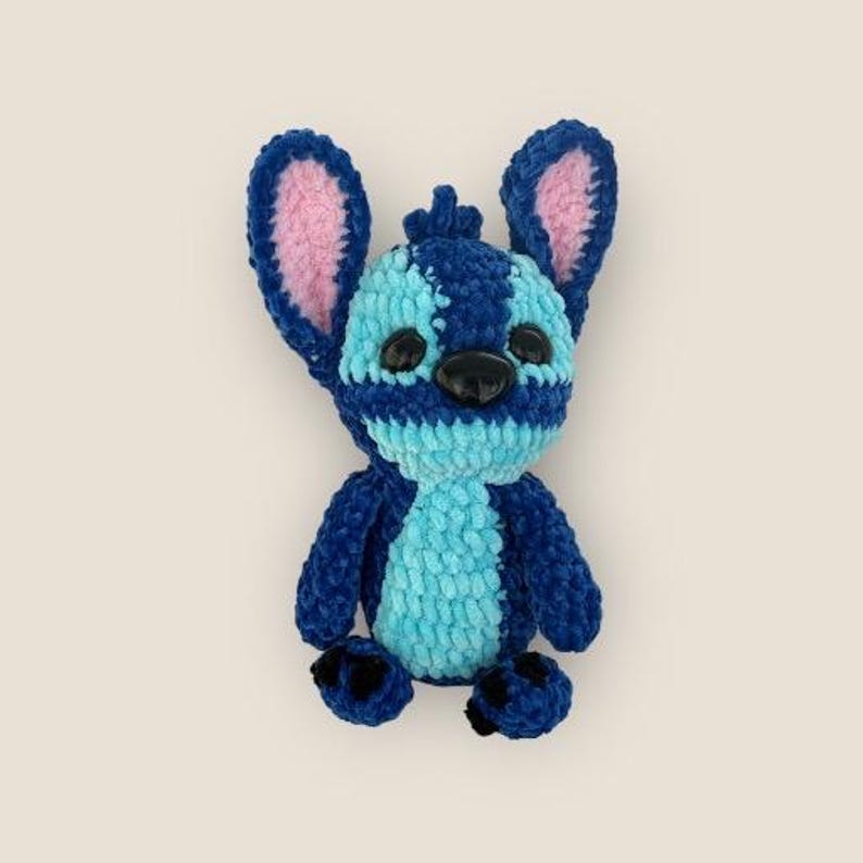 May include: A plush toy of a blue alien character with large ears and black eyes. The toy has a light blue body, dark blue stripes, and pink inner ears. The character's feet are dark blue with black paw pads.