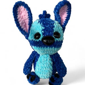 May include: A handmade plush toy in the shape of a blue alien creature with large ears and black eyes. The toy has a light blue chest and face, with darker blue stripes and accents. The inside of the ears is pink.