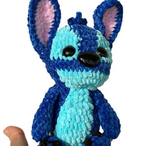 May include: A handmade blue and light blue crochet plush toy with large ears and black eyes. The plush toy is held in a hand against a white background. The toy's ears have pink inner linings.