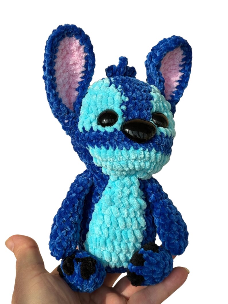 May include: A handmade blue and light blue crochet plush toy with large ears and black eyes. The plush toy is held in a hand against a white background. The toy's ears have pink inner linings.