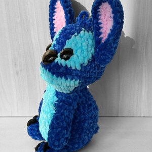 May include: A handmade blue crochet plush toy with large ears and black eyes. The plush features light blue and pink accents, and is sitting down. The toy is made of soft, textured yarn.