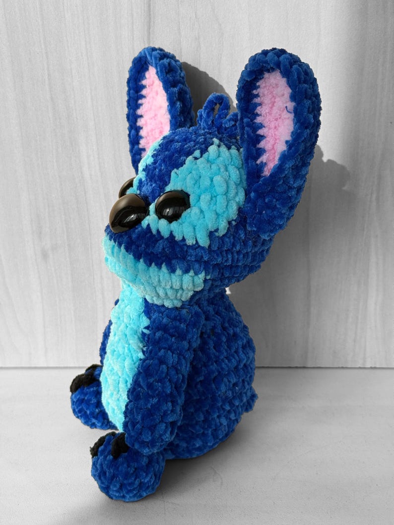 May include: A handmade blue crochet plush toy with large ears and black eyes. The plush features light blue and pink accents, and is sitting down. The toy is made of soft, textured yarn.