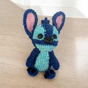 May include: A handmade plush toy of a cartoon character, featuring a blue and turquoise color scheme. The toy has large ears with pink inner details, black eyes, and a black nose. The plush is made of a soft, textured material.
