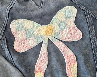 May include: A blue denim hoodie featuring a large patchwork bow appliqué. The bow is made from various fabrics in shades of pink, yellow, green, and white.