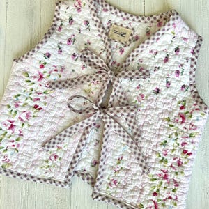 May include: A quilted vest featuring a floral pattern in pink and white, with a gingham trim and bow closure. The vest has a diamond-stitched pattern and a small tag with text.