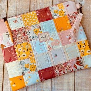May include: A patchwork zippered pouch with a wrist strap. The pouch features a variety of floral and geometric patterns in shades of yellow, pink, red, and blue. A small label with text is sewn onto the front. The zipper pull is a yellow tassel.