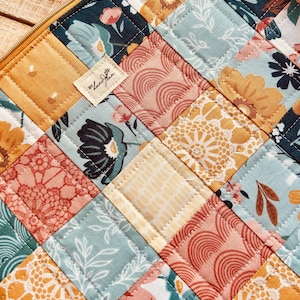 Puede incluir: Primer plano de una tela acolchada con un diseño de patchwork. Los cuadrados presentan estampados florales en tonos amarillos, turquesa, coral y azul marino. Una pequeña etiqueta con texto está cosida en la tela. Una cremallera dorada es visible.