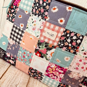 Puede incluir: Un estuche con cremallera de patchwork con una variedad de patrones florales y geométricos en rosa, azul, negro y blanco. El estuche tiene una cremallera marrón y una lengüeta de cuero. Una pequeña etiqueta dice "by lanijohn".